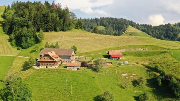 Een landhuis met een grote schuur, omgeven door groene velden en beboste heuvels.