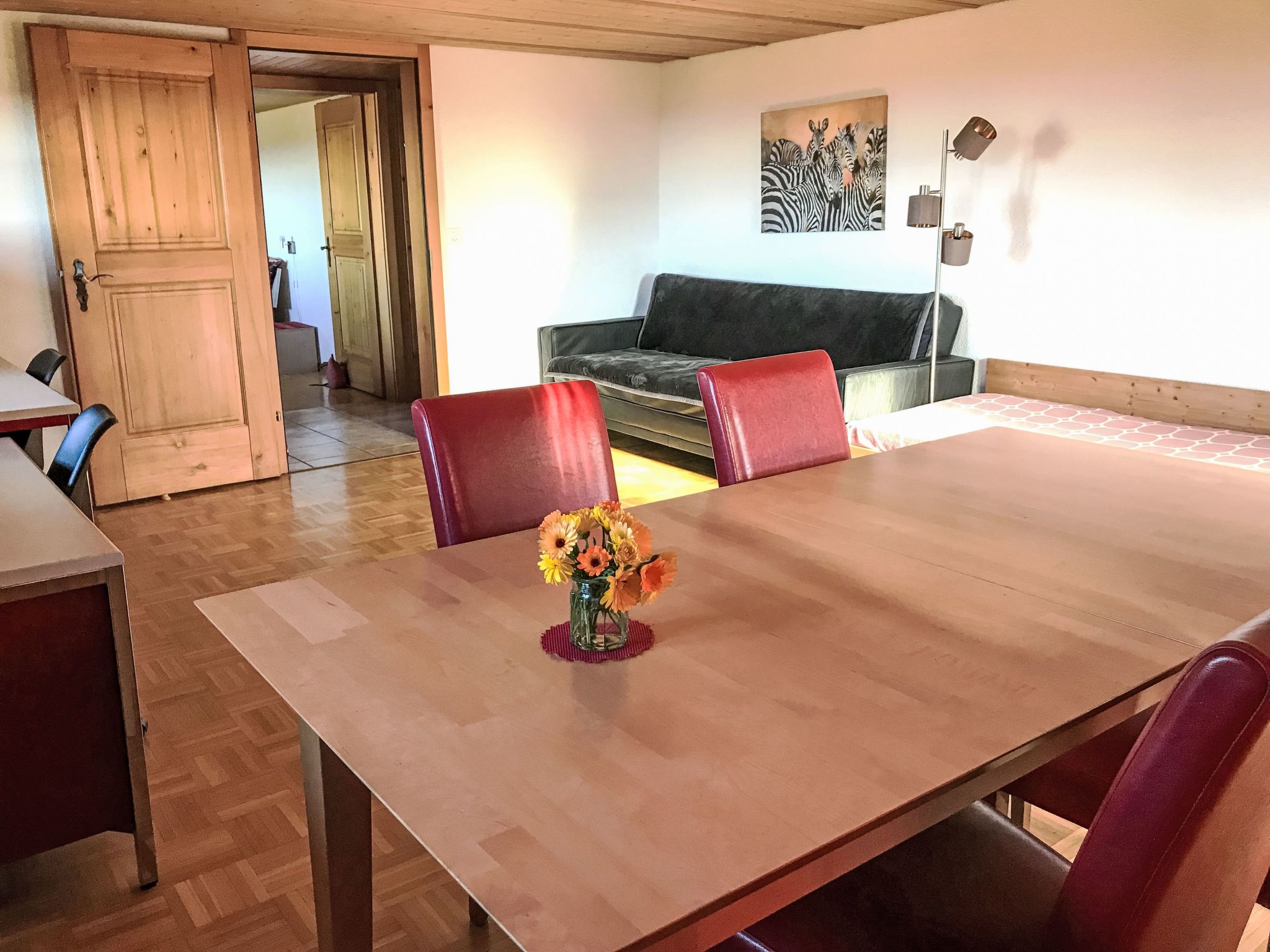 Een eetgedeelte met een houten tafel, rode stoelen en een bloemenvaas. Er is een bank en een schilderij aan de muur.