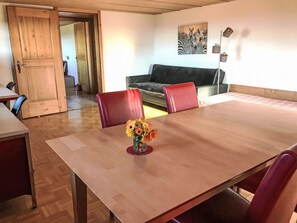 Ein Essbereich mit einem Holztisch, roten Stühlen und einer Blumenvase. Es gibt ein Sofa und ein Bild an der Wand.