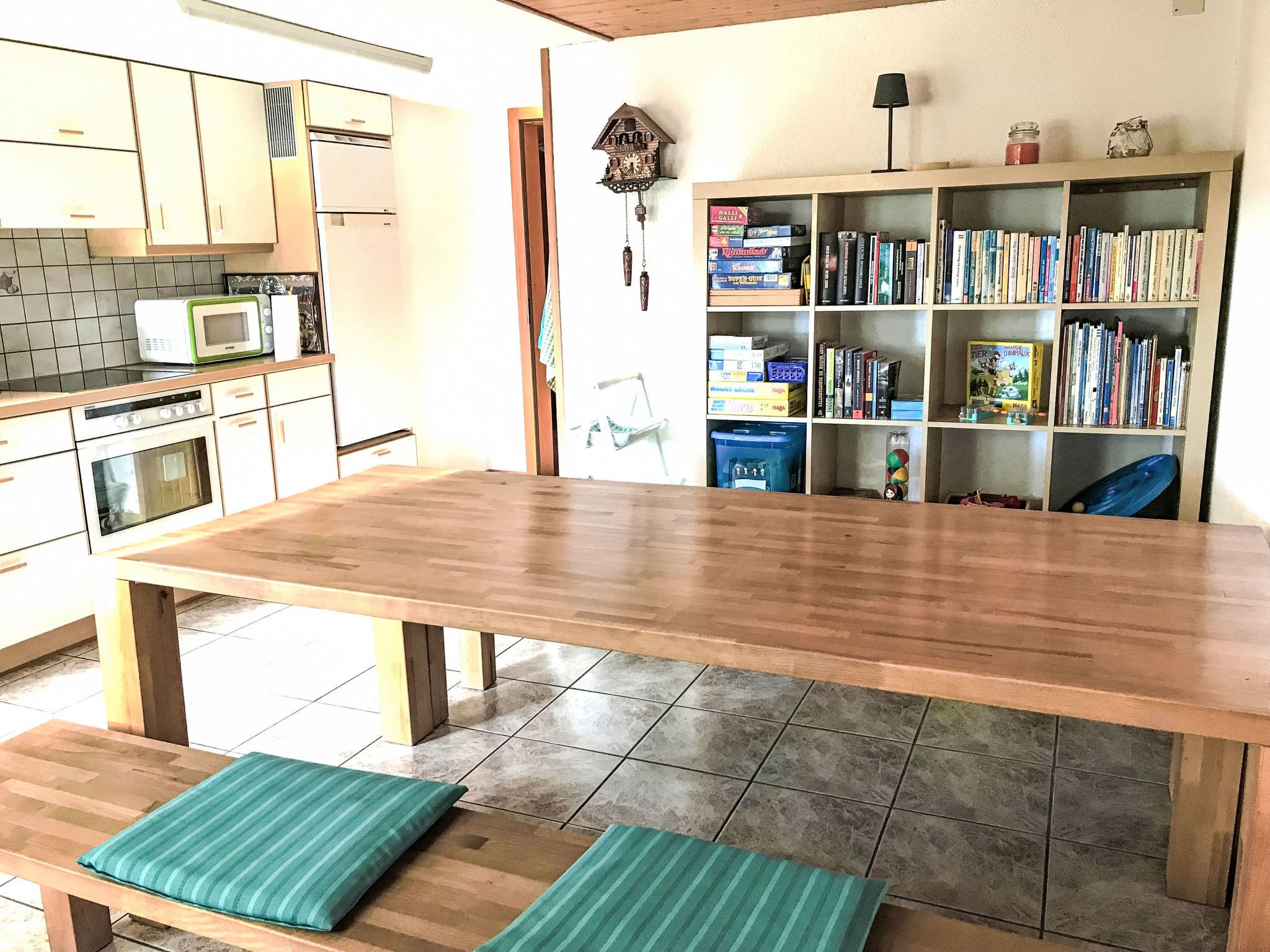 Een keuken met een houten tafel, bankjes, een magnetron en een boekenplank vol boeken.
