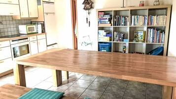 Een keuken met een houten tafel, bankjes, een magnetron en een boekenplank vol boeken.