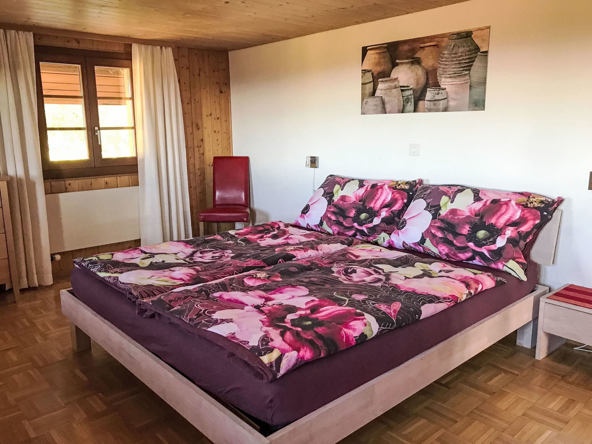 Un dormitorio con cama, una silla, un cuadro en la pared y un ventanal con cortinas.