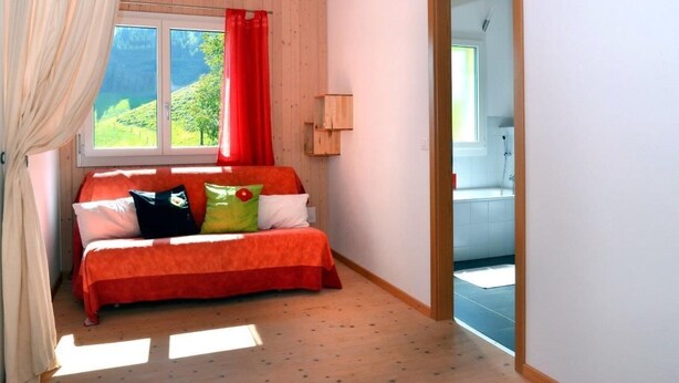 Um quarto com cama, janela com vista para vegetação, piso de madeira e porta que dá para um banheiro.