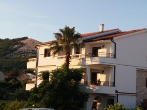 Un edificio a due piani con balconi, immerso nel verde e con una collina sullo sfondo.