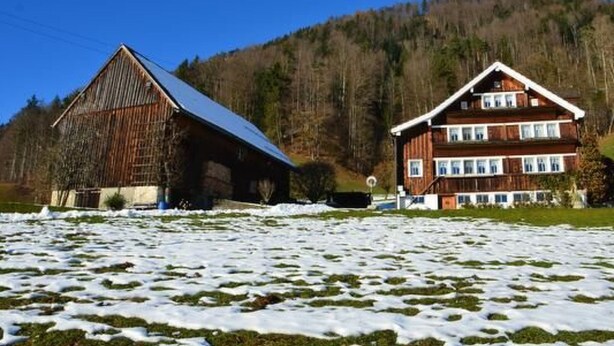 Una casa de madera de dos pisos con techo inclinado, rodeada por un paisaje nevado y una colina boscosa.