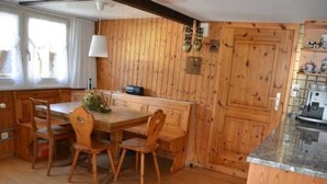 Un interior de madera con una mesa y sillas de comedor, una zona de cocina con armarios de madera y una ventana con cortinas.