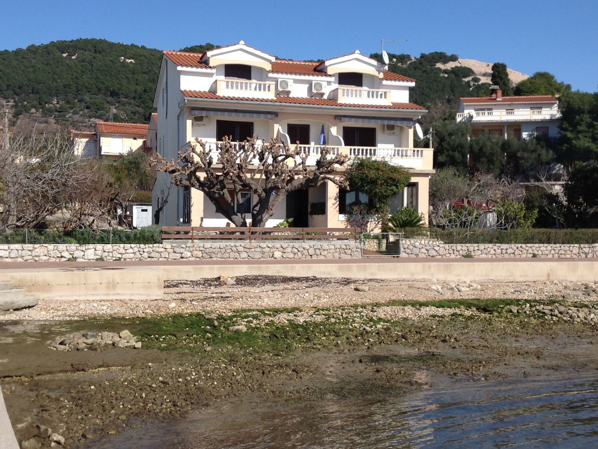 Uma casa de dois andares com uma varanda, um muro de pedra na frente e uma árvore em primeiro plano.