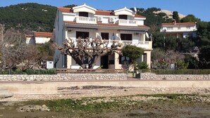 Una casa a due piani con un balcone, un muro in pietra di fronte e un albero sullo sfondo.