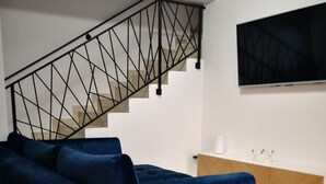 En moderne stue med en blå modulsofa, en trappe med geometrisk gelænder, et træskab og et fladskærms-tv monteret på væggen.