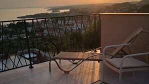En balkon med to fletstole og et lille bord, der ud til et smukt udsigtspunkt over vand og fjerne bakker i solnedgangen.