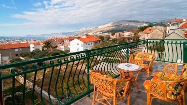 Un balcone con sedie in vimini e un tavolo, con vista su una cittadina costiera con edifici a tetti rossi e una montagna sullo sfondo.
