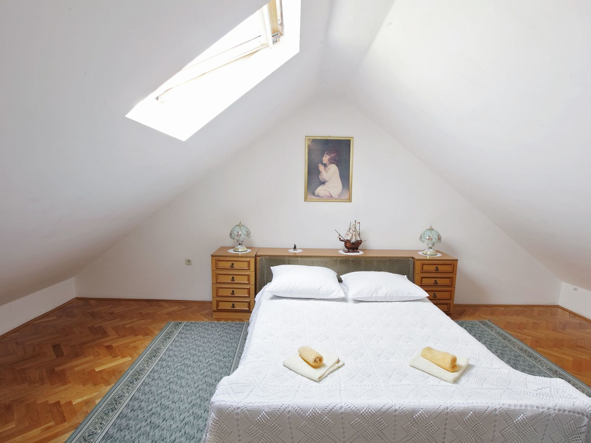 A bedroom with a sloped ceiling, a bed with two pillows, a nightstand with a lamp, and a painting on the wall.