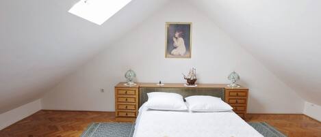 A bedroom with a sloped ceiling, a bed with two pillows, a nightstand with a lamp, and a painting on the wall.