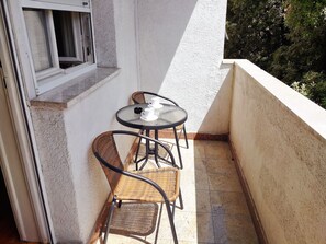 Een klein balkon met een tafel en twee rieten stoelen, een raam en een witte muur.