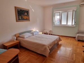 Een slaapkamer met een bed, nachtkastjes, een klein tafeltje, een raam met jaloezieën en een schilderij aan de muur.