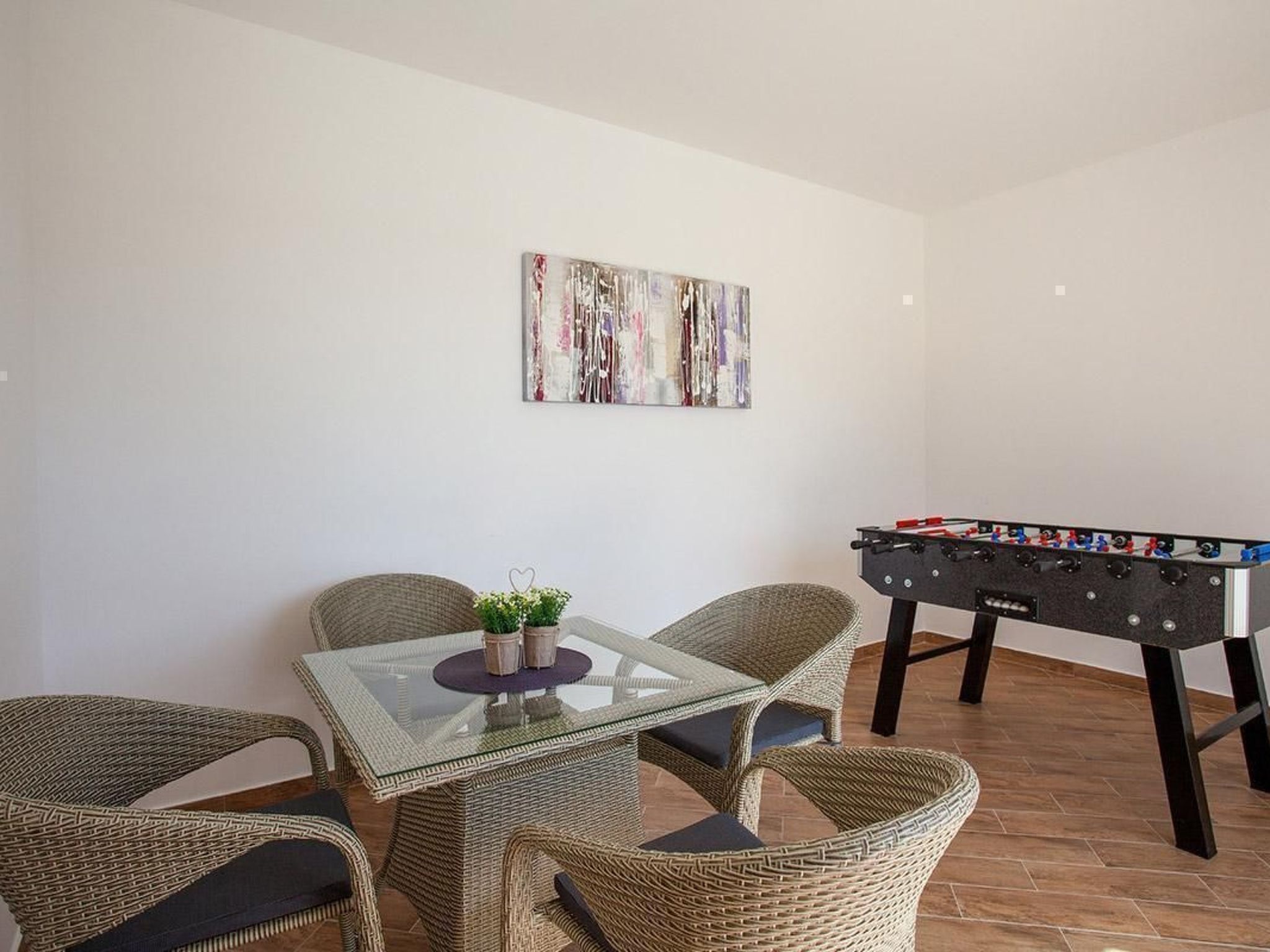 Une pièce avec une table dont le plateau est en verre, entourée de chaises en osier, une table de baby-foot et une œuvre d’art abstraite accrochée au mur.