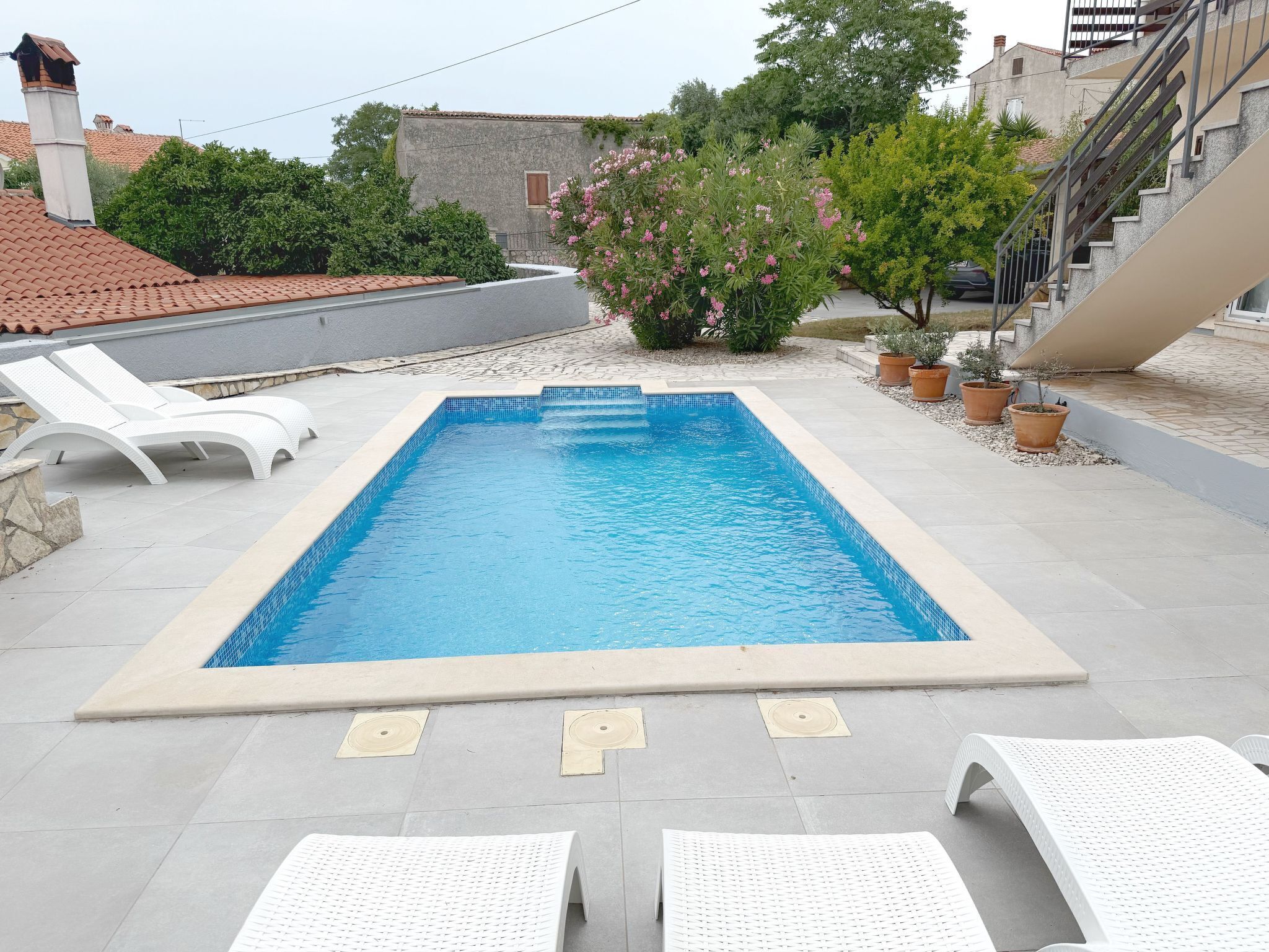A rectangular swimming pool with clear blue water, surrounded by white lounge chairs and potted plants.