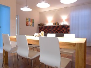 Un comedor con una mesa de madera y sillas blancas, iluminación colgante y una pared a cuadros detrás de la mesa.
