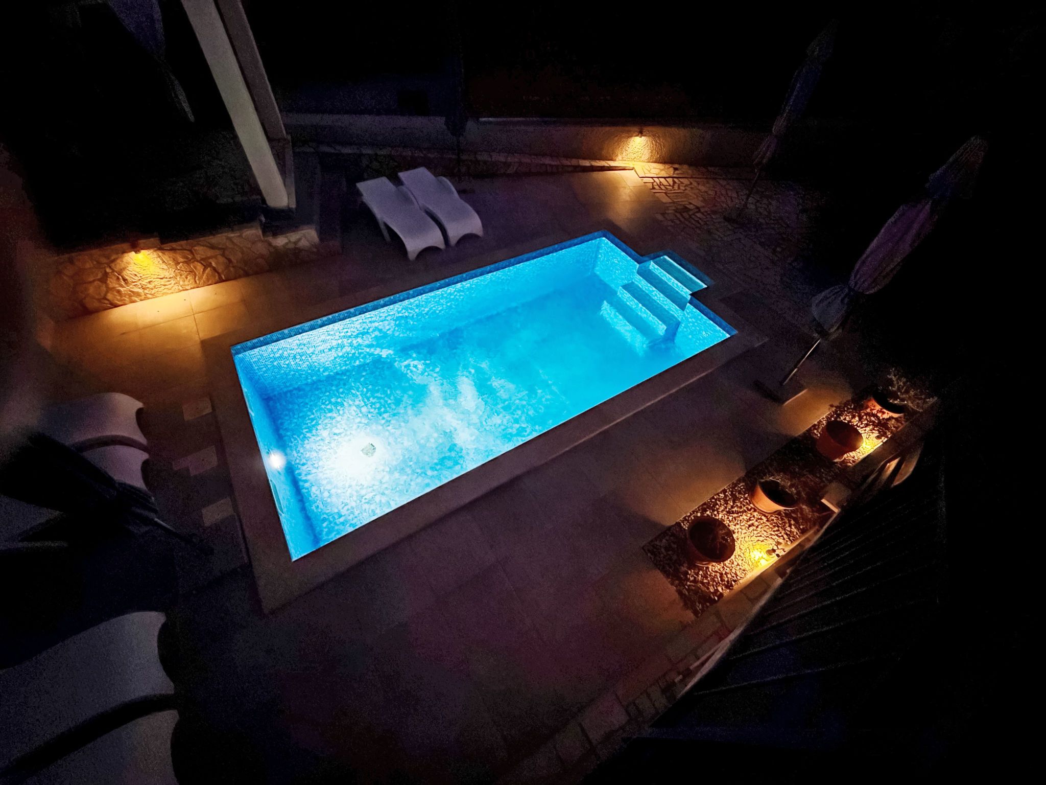 A rectangular swimming pool with a blue illuminated interior, surrounded by stone tiles and a low wall.