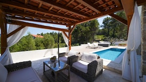 Un espace extérieur aménagé, abrité et doté de meubles en osier et d’une table en verre, offrant une vue sur une piscine et un jardin luxuriant.