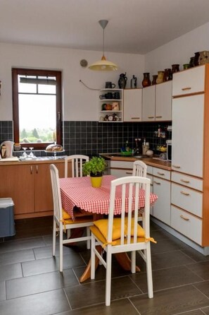 A compact kitchen with white cabinets, a tiled backsplash, a small dining table with a checkered tablecloth, and a single chair.
