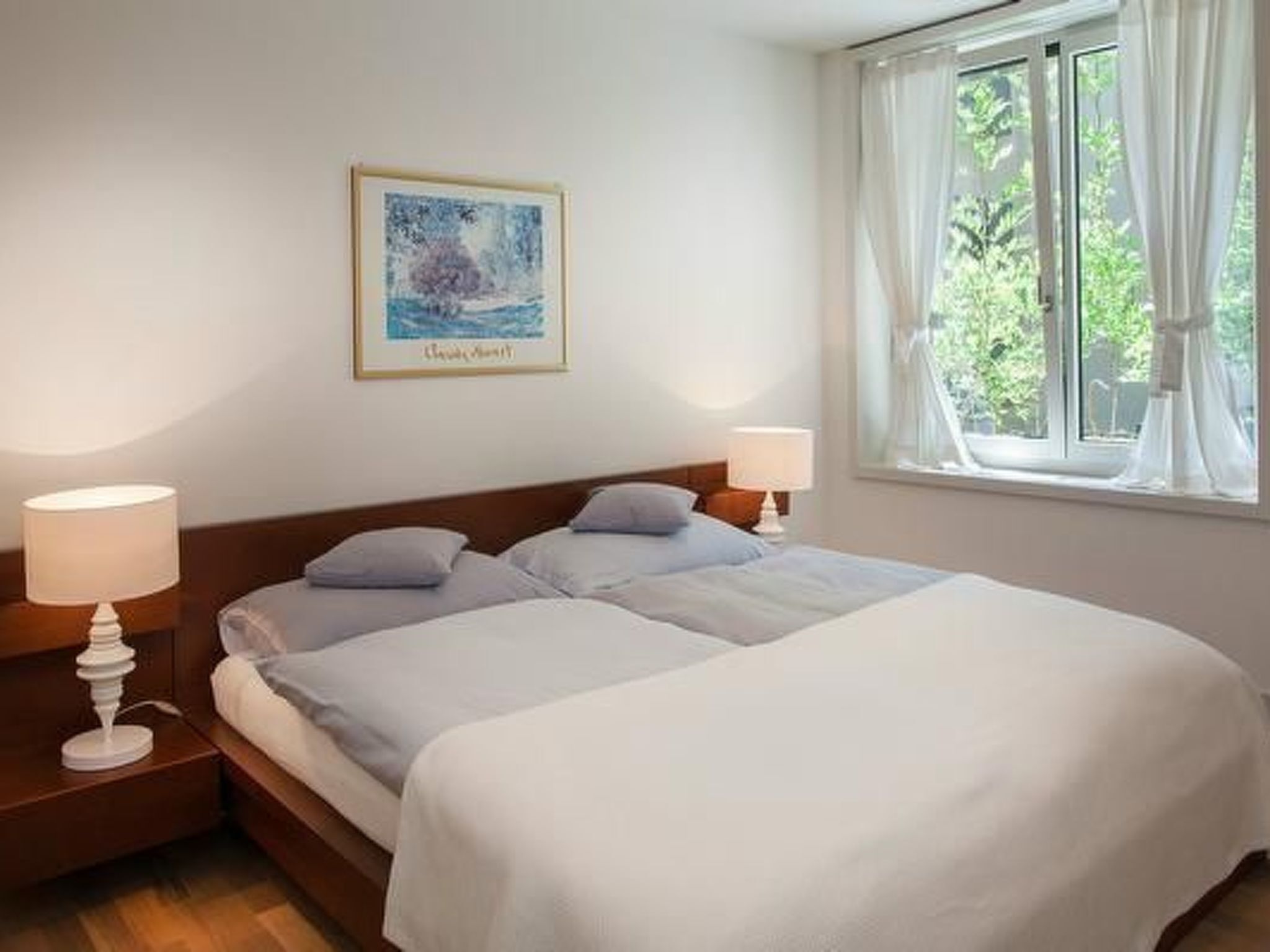A double bed with white linens, two bedside lamps, a framed artwork on the wall, and a window with curtains.