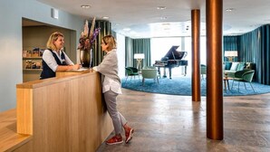 A hotel reception area with a wooden reception desk, a piano in the background, and a spacious lobby with a high ceiling and large columns. - Parkresidenz Meilen by Interhome (Meilen)
