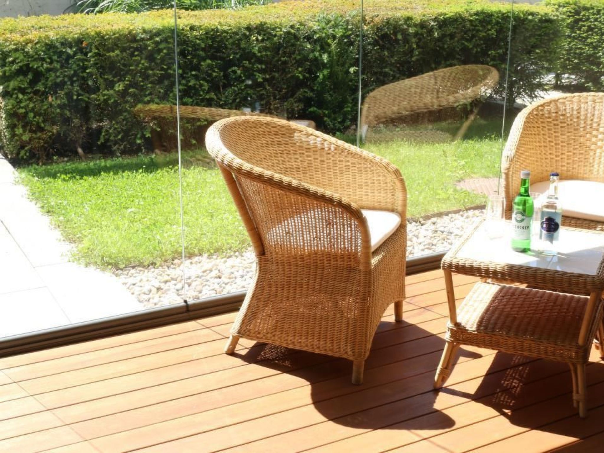 Une chaise et une table en osier, sur une terrasse en bois dotée d’une balustrade en verre.