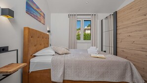 A modern bedroom with a wooden headboard, a bedside table, a window with curtains, and a view of greenery outside. - Villa Lejla by Interhome (Drenje)