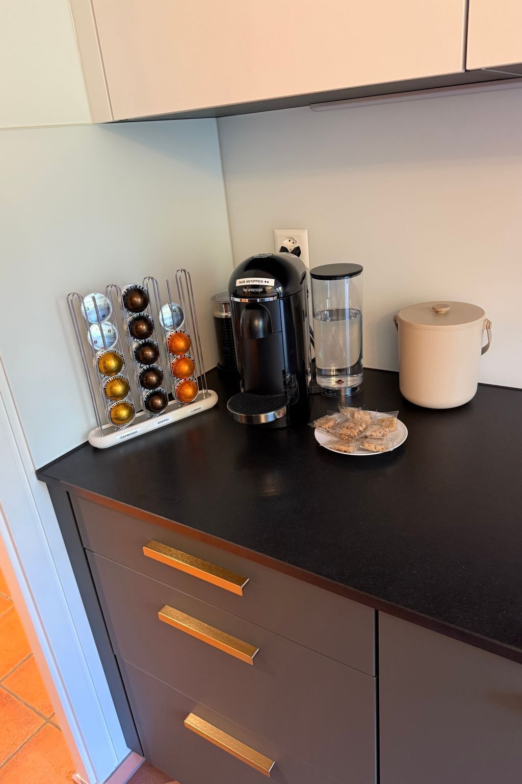 Una encimera de cocina con una cafetera, un dispensador de agua, un vaso de agua, un plato de galletas y un soporte para cápsulas de café.