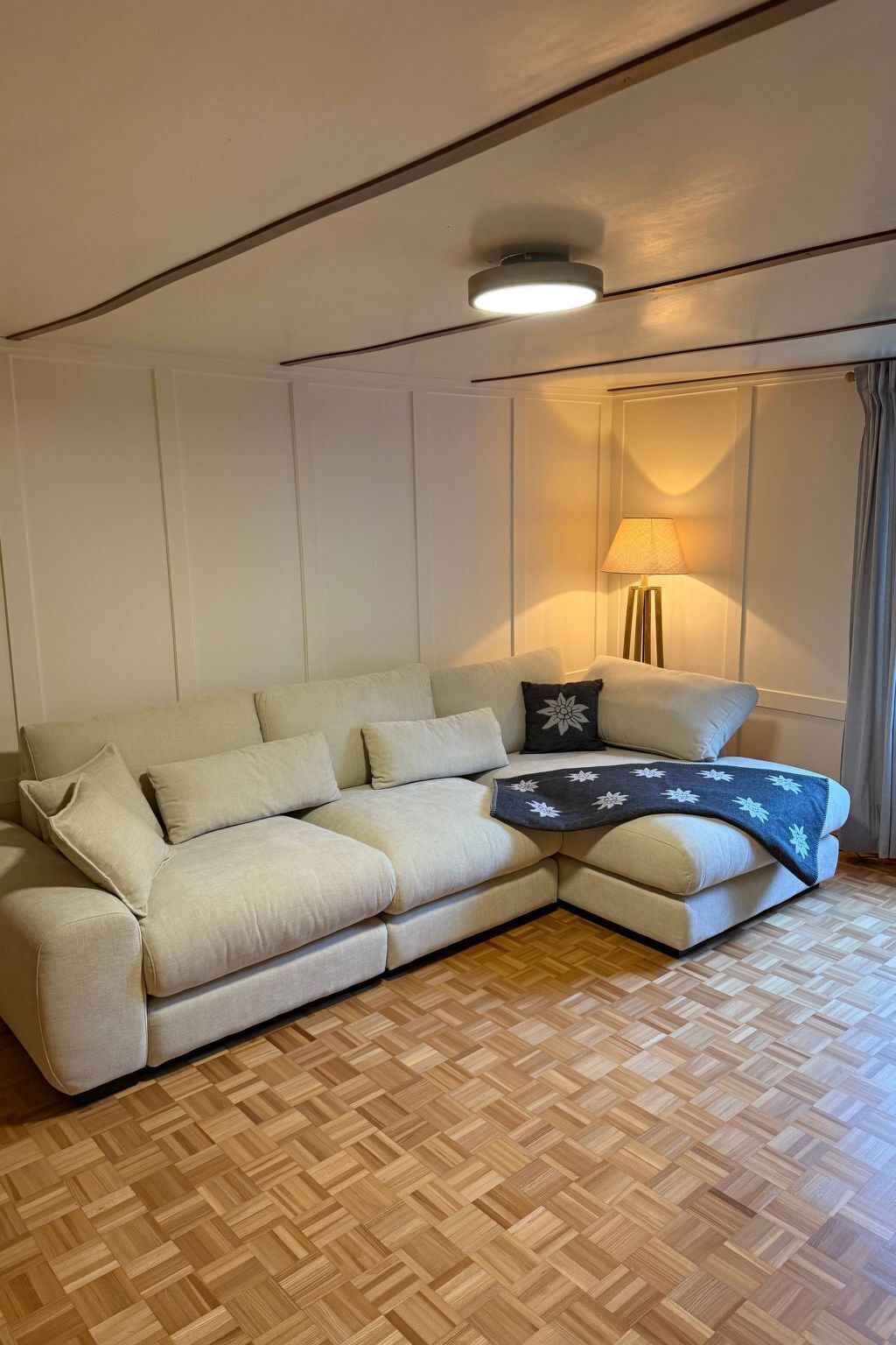 A living room with a beige sectional sofa, a patterned throw blanket, a floor lamp, and a wooden parquet floor.