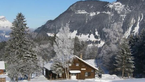 A wooden cabin with a snow-covered roof, surrounded by snow-covered trees and mountains in the background. - Ferien im grünen links by Interhome (Haldi b. Schattdorf)