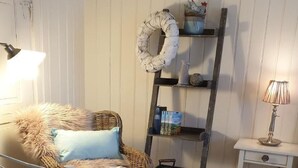 Una habitación acogedora con una silla de mimbre, una cama blanca, una estantería de madera escalonada y una mesita de noche con una lámpara.