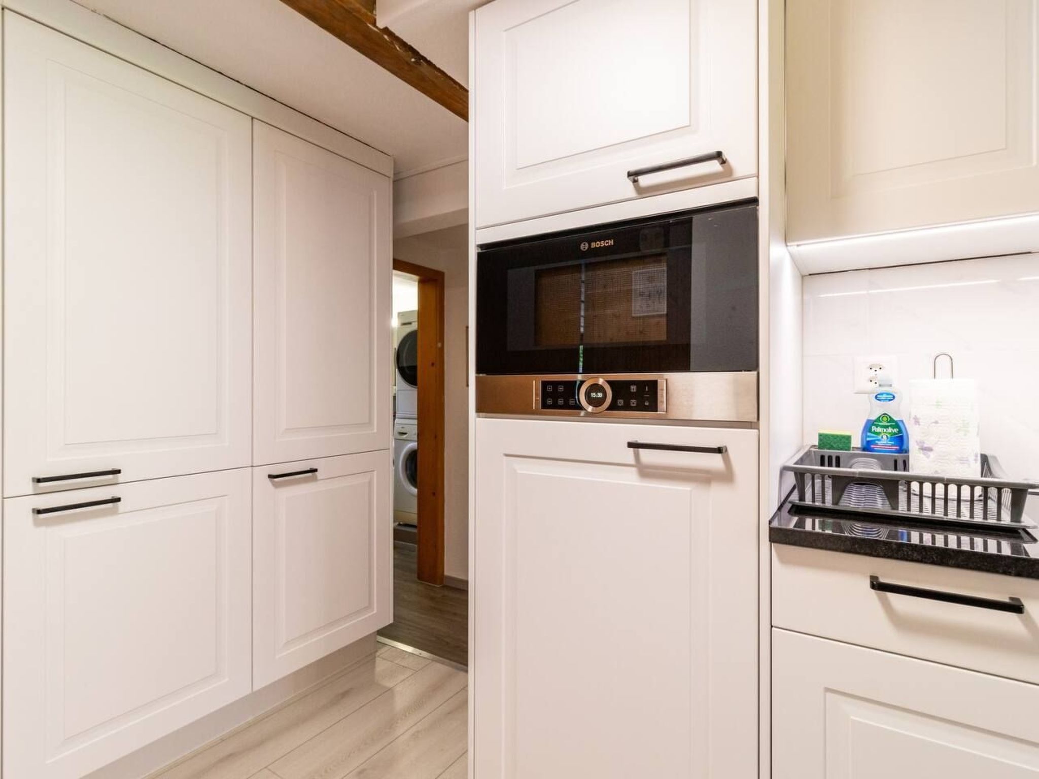 Une cuisine avec des meubles blancs, un four à micro-ondes de la marque Bosch et un égouttoir à vaisselle contenant des produits de nettoyage.