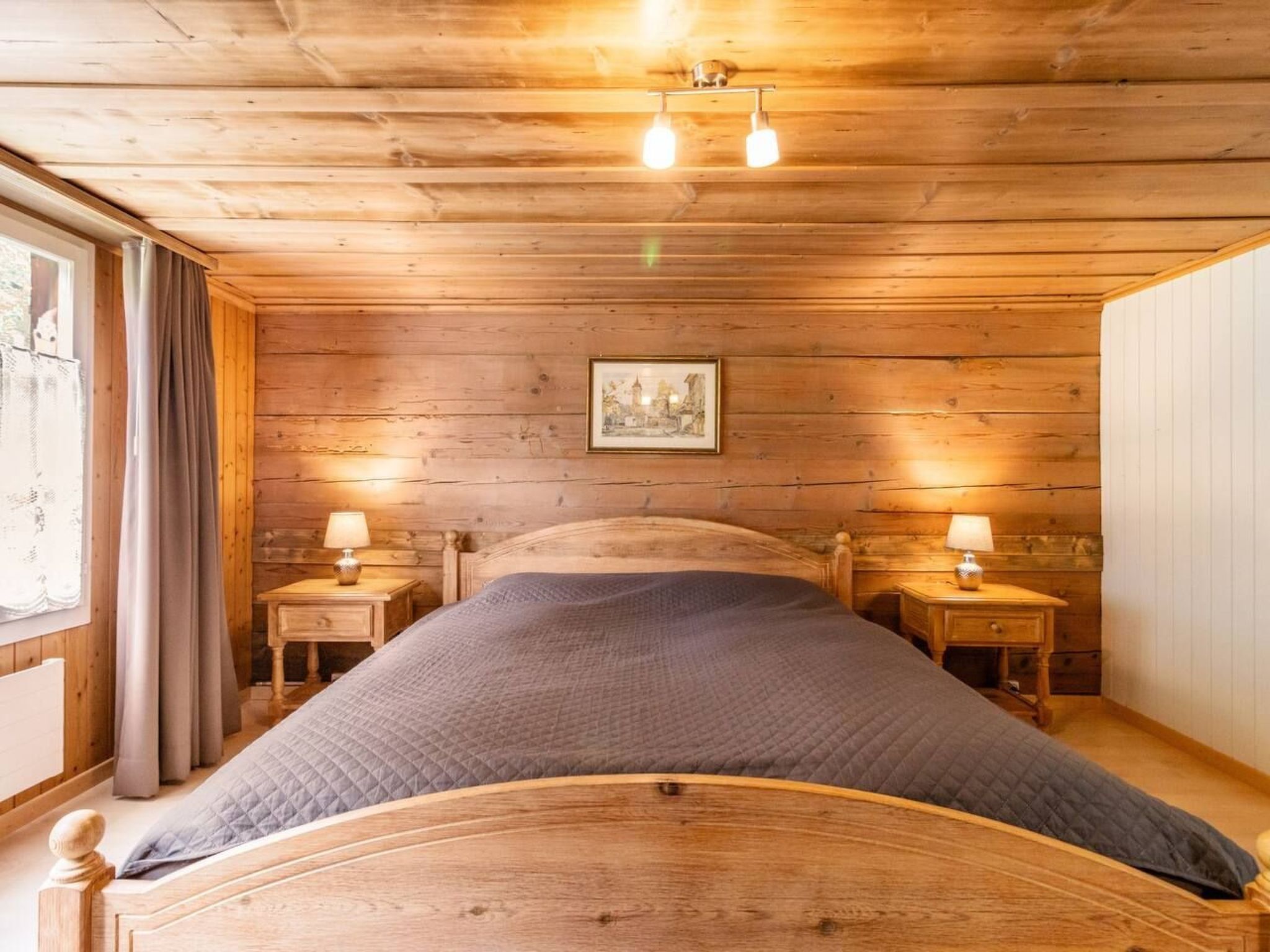 Ein schlichtes Schlafzimmer im Blockhausstil mit einem großen Bett, zwei Nachttischen mit Lampen, einem gerahmten Bild an der Wand und einem Fenster mit Vorhängen.