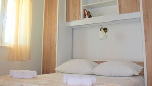 Um quarto com cama, duas almofadas, mesa de cabeceira, estante com livros e um pequeno vaso, e janela com cortinas.