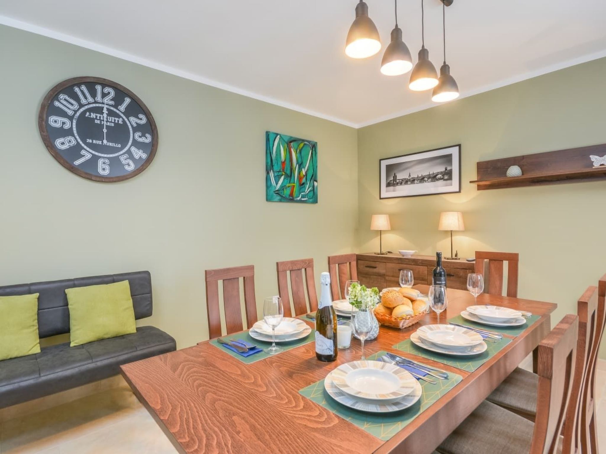 A dining area with a wooden table set for four, surrounded by chairs. A large wall clock and a framed picture are visible.