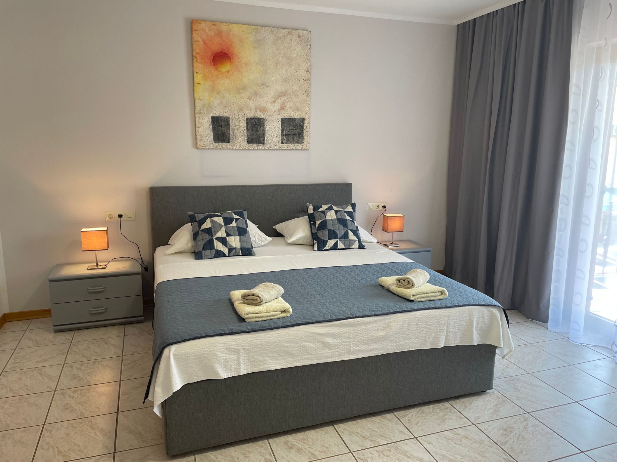 Una cama bien hecha con una cubierta azul acolchada, dos almohadas y dos toallas dobladas. Hay una mesita de noche con una lámpara y un enchufe. Un cuadro cuelga sobre la cama.