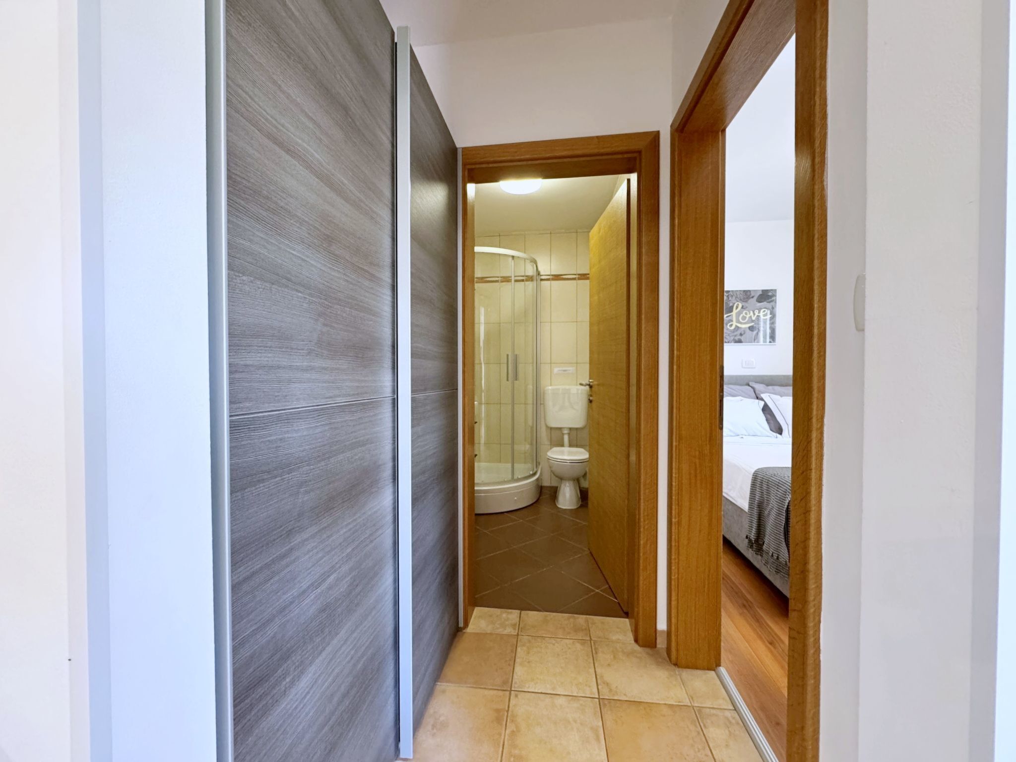 A narrow hallway leading to a bathroom with a shower, toilet, and a bed visible through an open door.