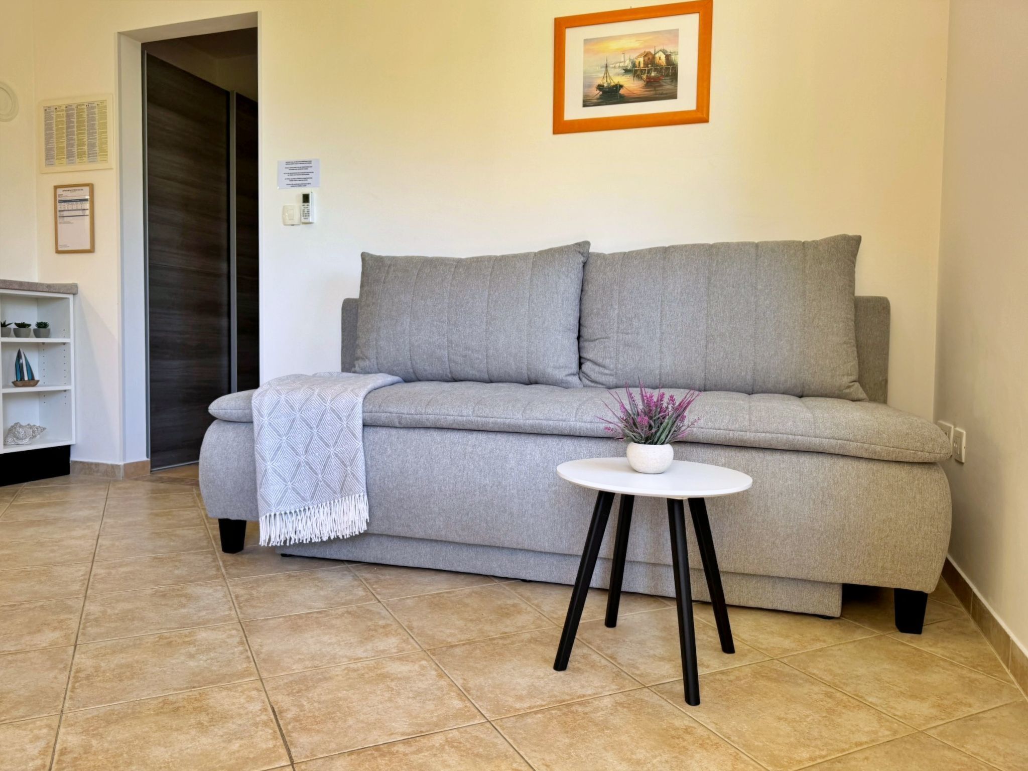 Ein modernes Wohnzimmer mit einem grauen Sofa, einem kleinen weißen Tisch mit einer Topfpflanze und einer Tür zu einem anderen Raum.