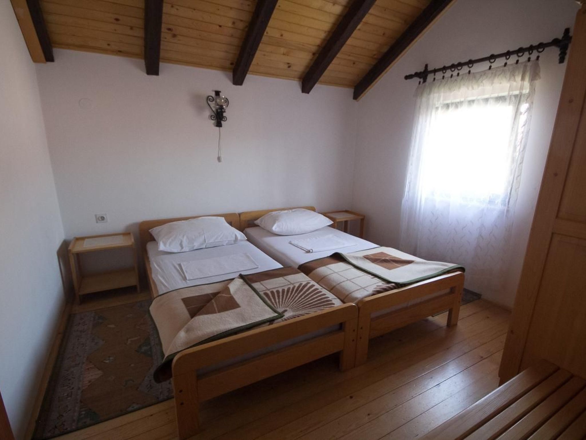 Ein Doppelbett mit Holzkopf- und Fußteil, zwei Nachttische, eine Wandleuchte und ein Fenster mit transparenten Vorhängen.