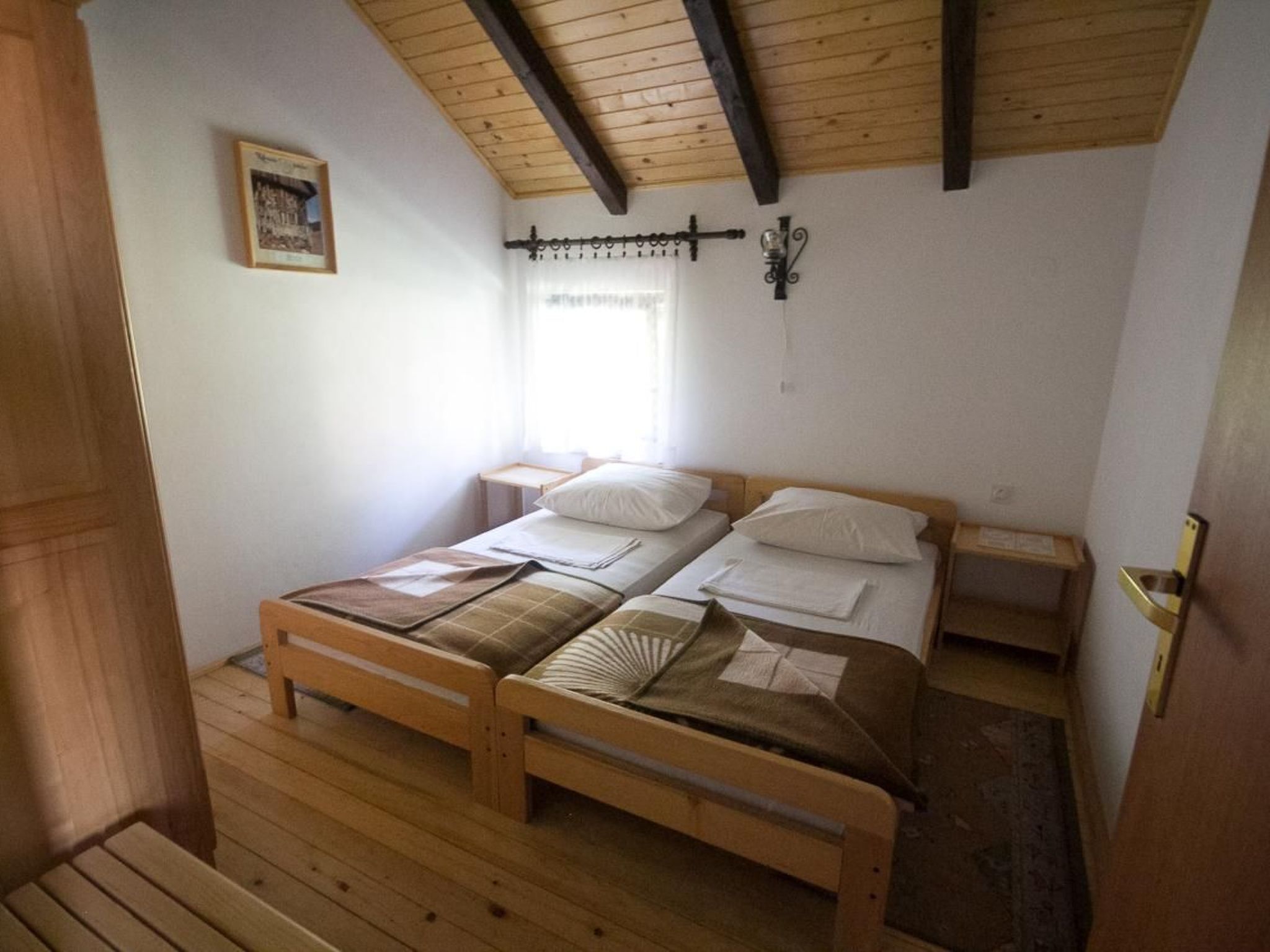 A small, cozy room with a wooden floor and a sloped ceiling. There are two single beds with wooden frames and white bedding. A small table and a wall-mounted light fixture are also visible.