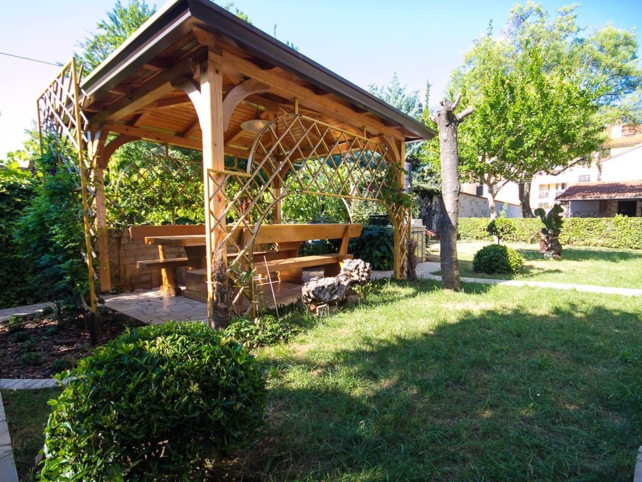 A wooden gazebo with lattice sides and a bench inside, surrounded by well-maintained grass and bushes.