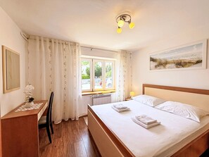 A bedroom with a bed, a desk, a chair, a window with curtains, a radiator, and a painting on the wall. - Apartment Nevenka by Interhome (Krk/Čižići)