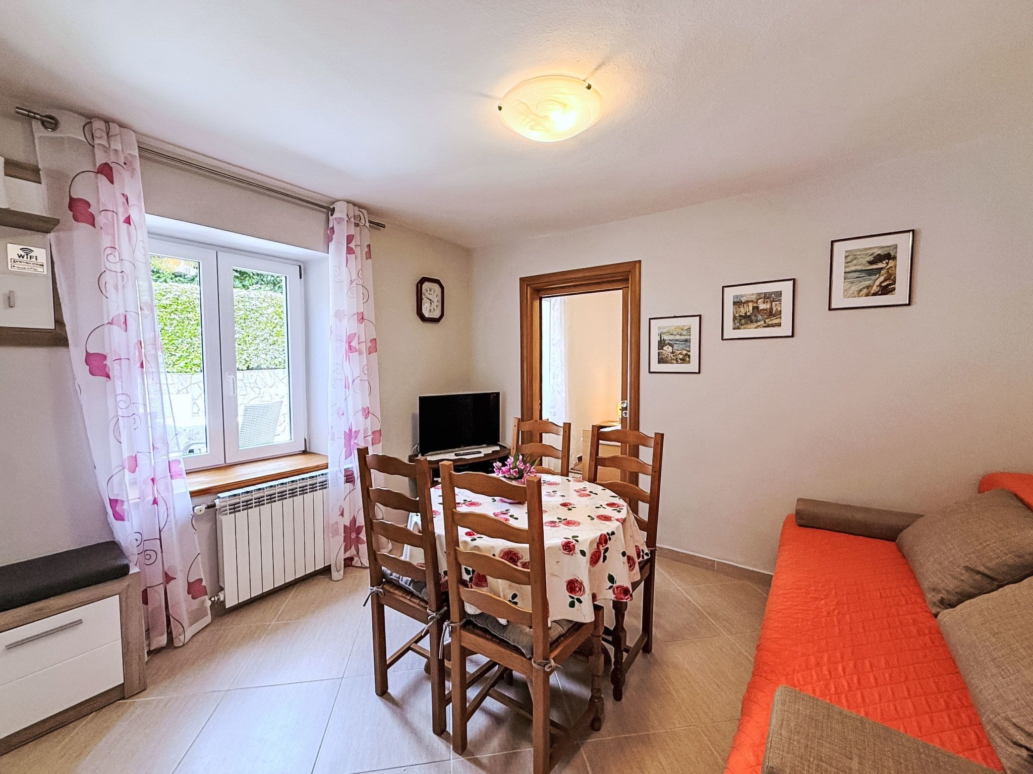 A room with a dining table, chairs, a TV, a bed, a window with curtains, a radiator, and framed pictures on the wall.