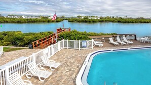 A pool area with white lounge chairs, a flag, and a view of a body of water. - Sea Shell Pool Townhouse Across from Beach (Indian Shores)