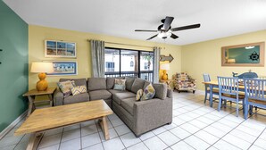 A living room with a sectional sofa, a wooden coffee table, a dining table with blue chairs, and a ceiling fan. - Sea Shell Pool Townhouse Across from Beach (Indian Shores)