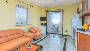 Sala de estar com sofá, poltrona e uma mesinha de centro com uma planta. Há uma televisão em um móvel, uma varanda com uma cadeira e uma área de cozinha com fogão e armários.