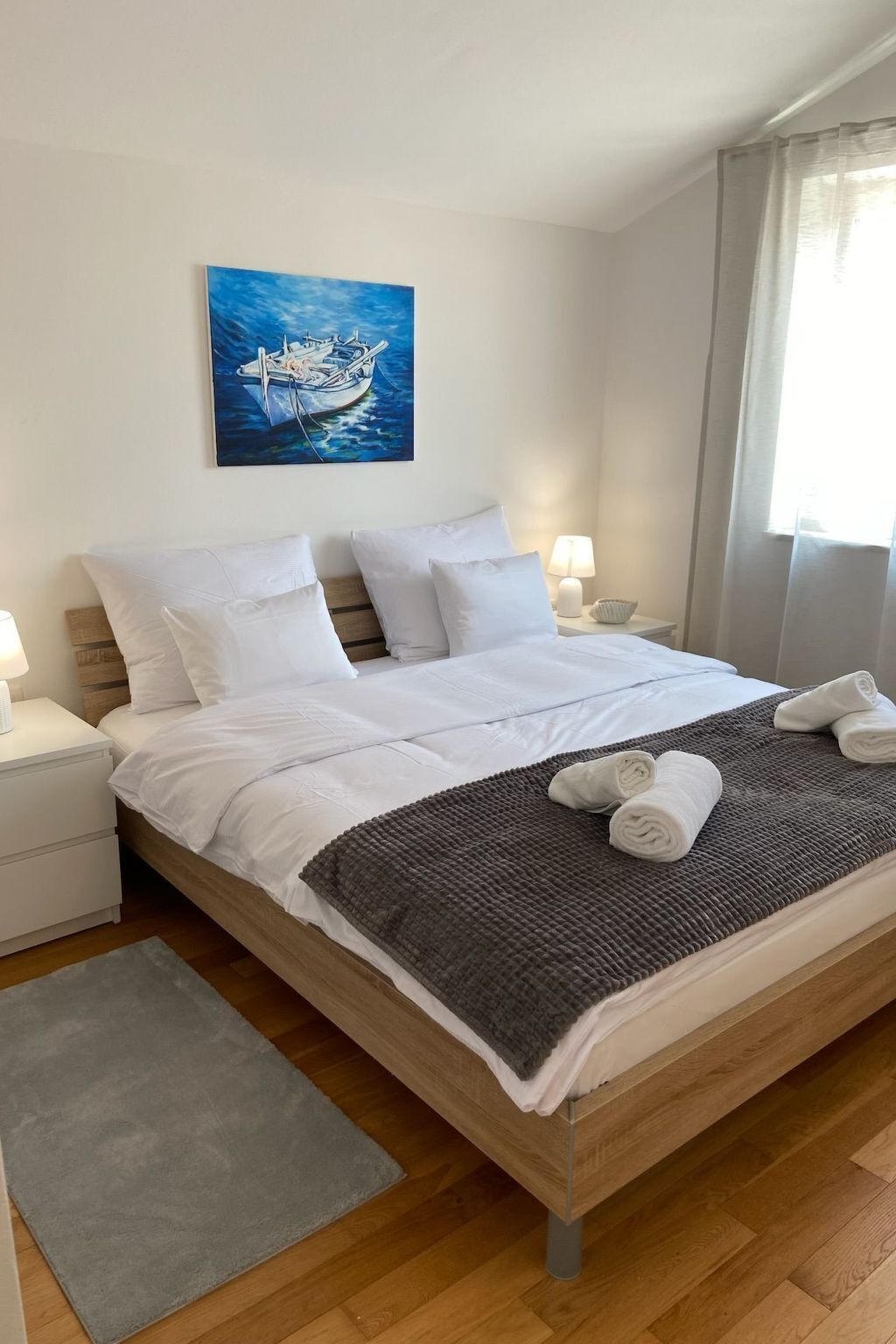 Una cama bien hecha con sábanas blancas y una manta gris, un cabecero de madera y un cuadro de un barco en la pared.