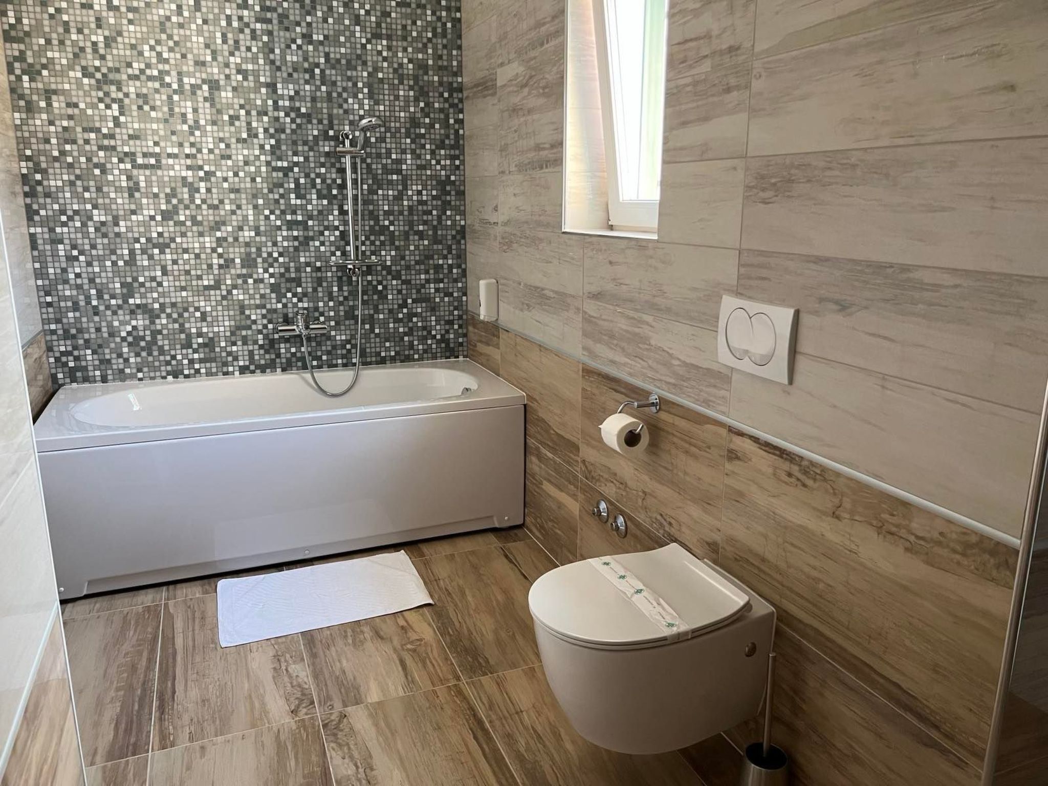 A modern bathroom with a mosaic tile shower wall, a freestanding bathtub, a toilet, and a window providing natural light.